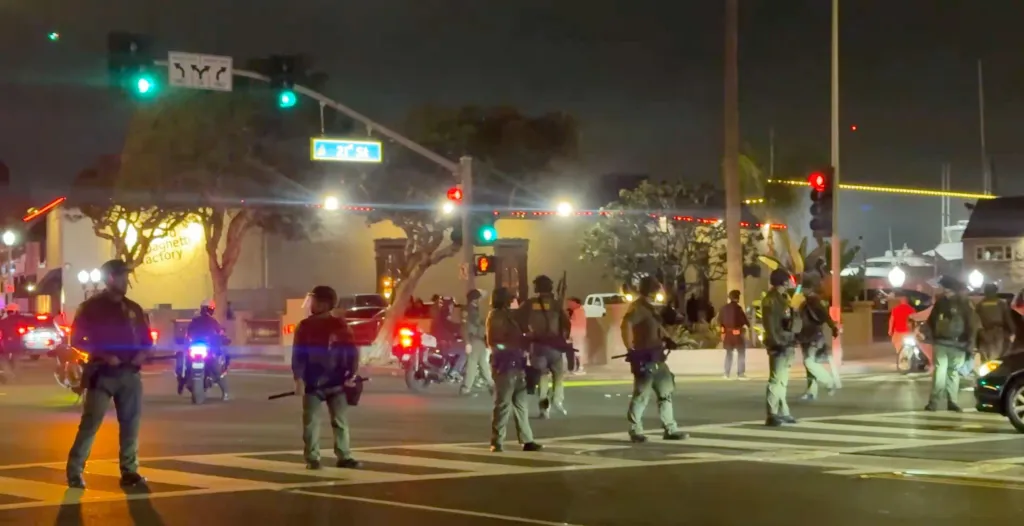 Law enforcement officers in riot gear stand in a street at night with traffic lights and buildings in the background.