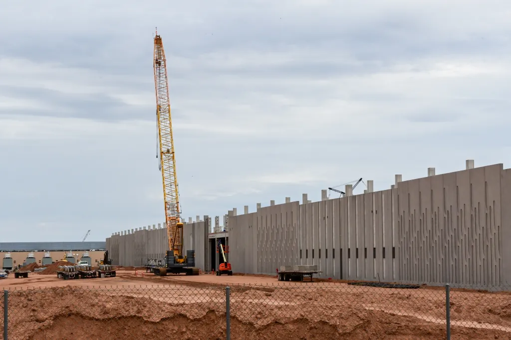 Construction of a data center in Mesa, Arizona, with a large crane and partially built gray concrete walls, seen from behind a chain-link fence.
