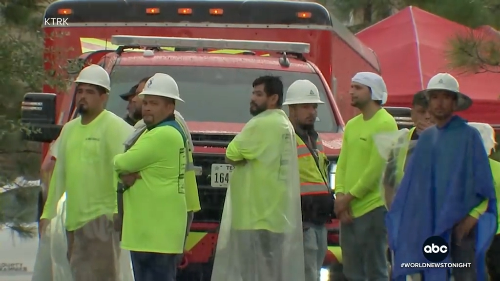 Construction workers in hard hats and neon shirts stand in front of a red emergency vehicle.