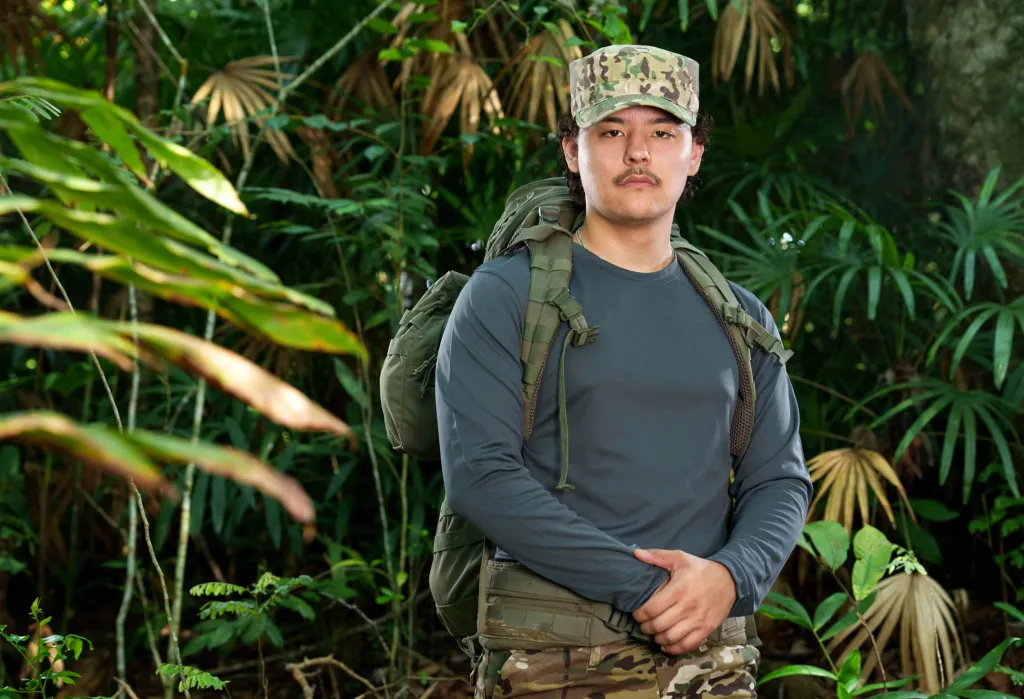 Collin Gosselin wearing a camo hat, dark grey shirt, camo pants, and a green backpack in a jungle setting.