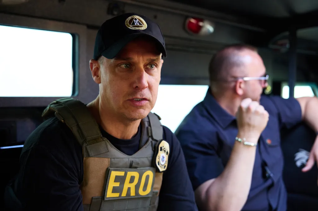 An agent wearing a tactical vest with "ERO" written on it and a black cap, sits next to a man in a dark blue shirt.