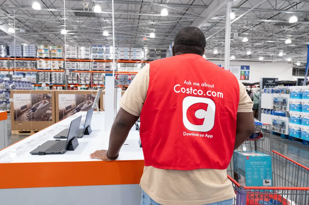 An employee in a red vest that reads "Ask me about Costco.com" and "Download our App" stands inside a Costco store.