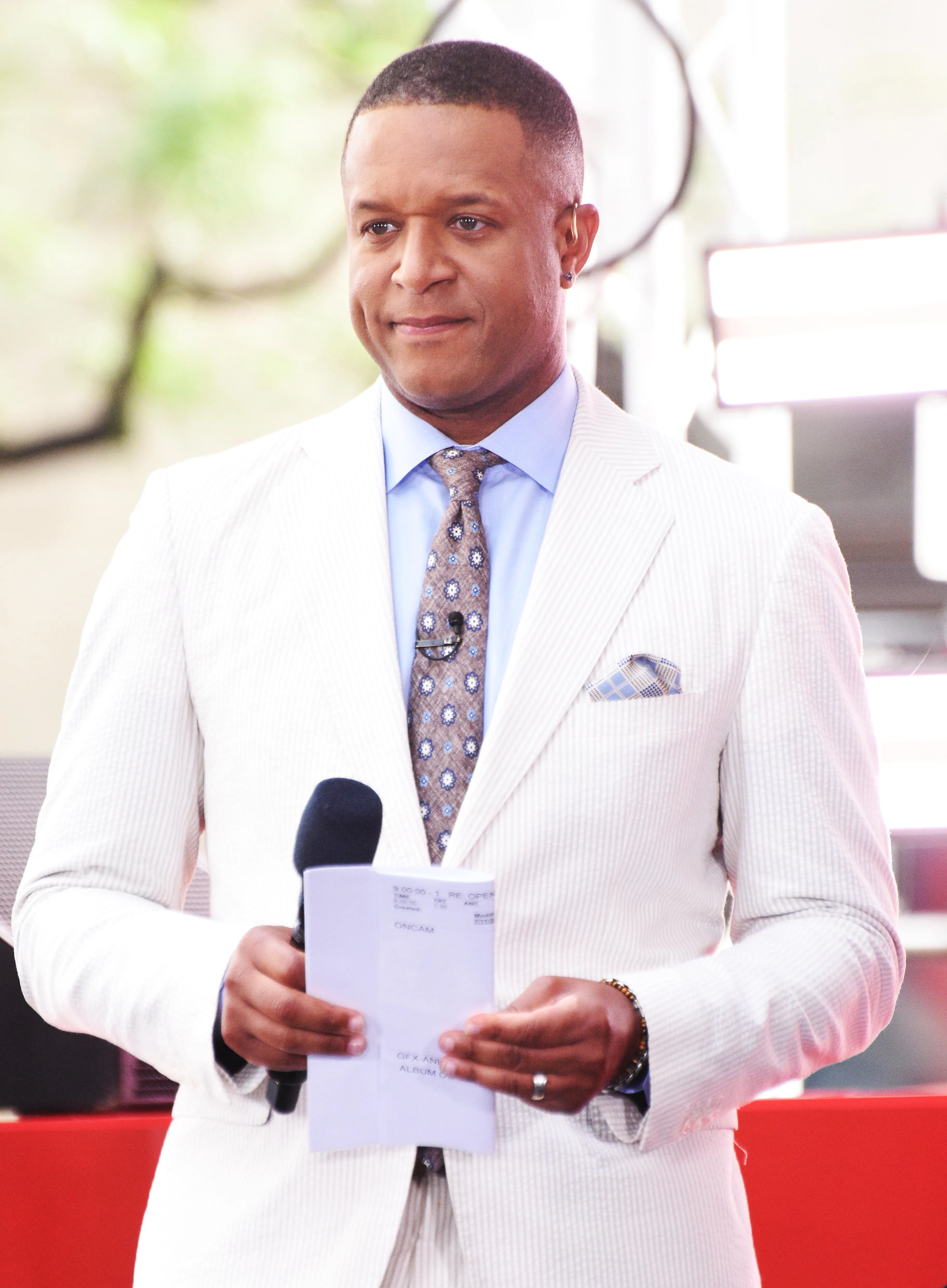 Craig Melvin wearing a white suit, light blue shirt, and patterned tie, holding a microphone and papers with "ONCAM" visible.