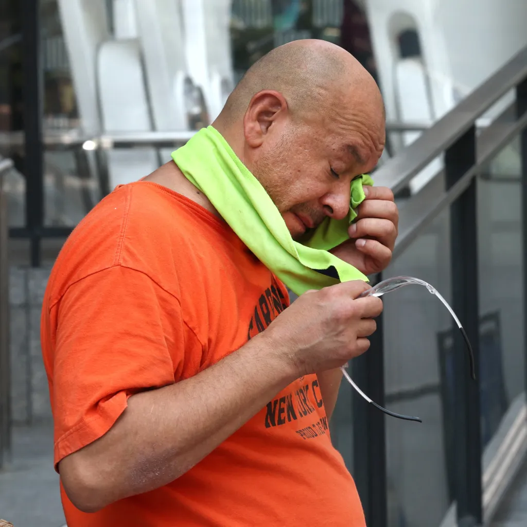 A man wiping his sweaty face with a green cloth while holding safety glasses.