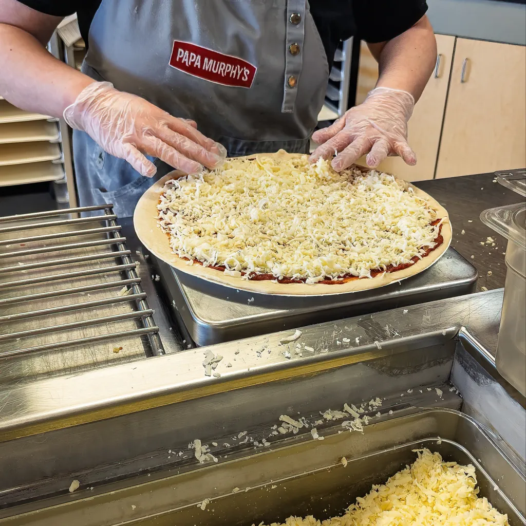 A Papa Murphy's employee preparing a pizza, sprinkling shredded cheese onto the dough.