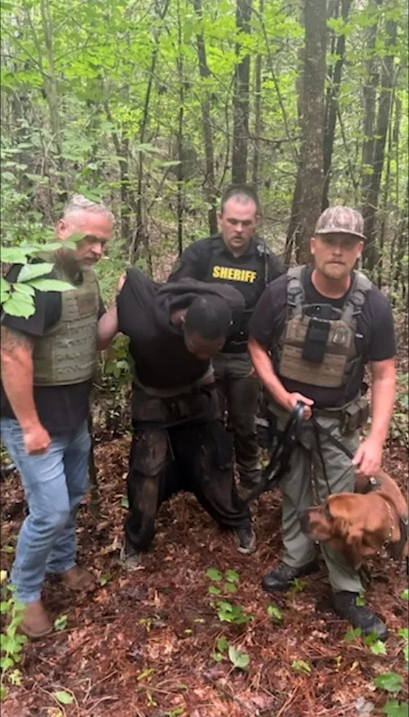 Darian Berry Sr. in custody, held by two officers, with a sheriff and a K-9 officer with his bloodhound nearby in a wooded area.