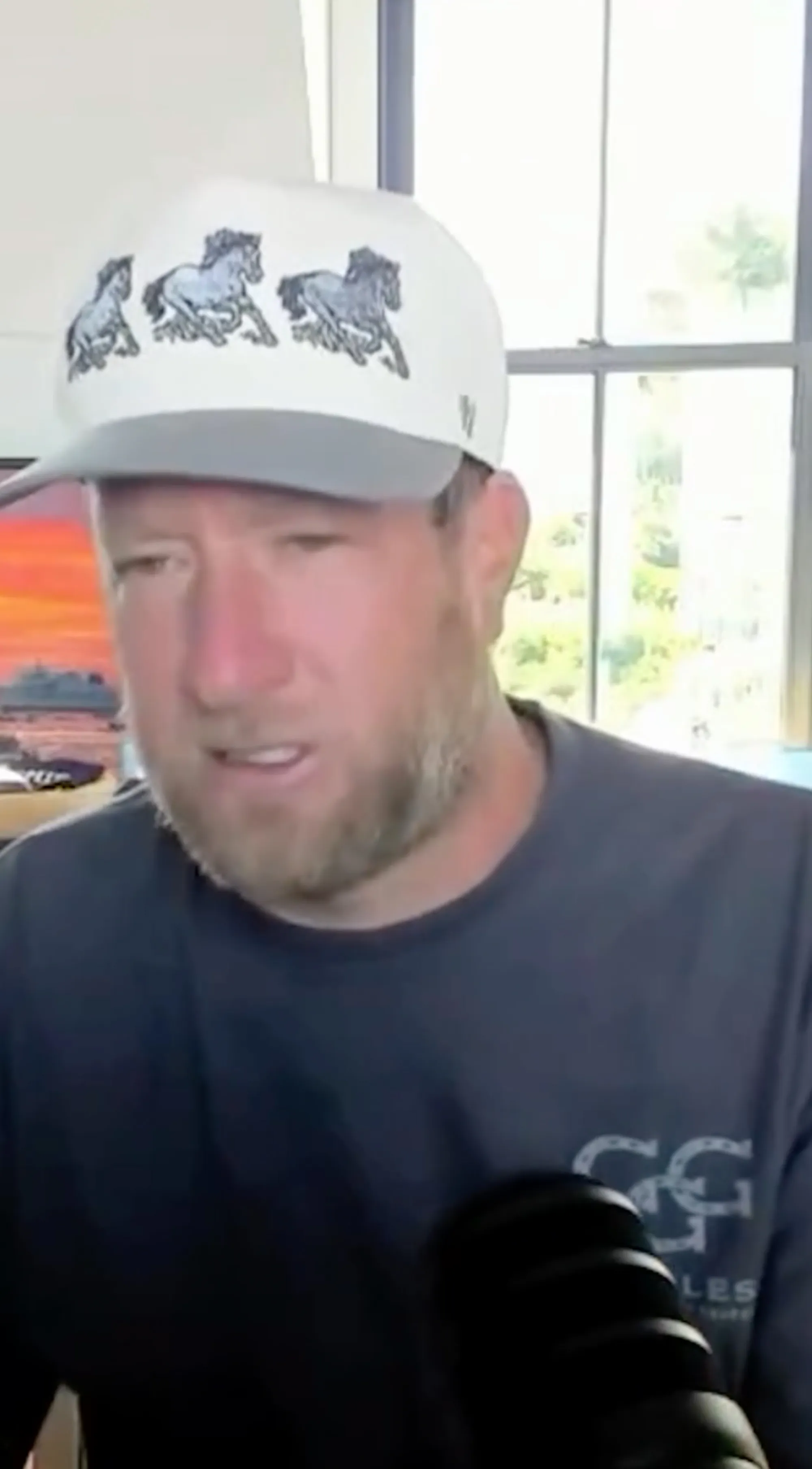 Dave Portnoy wearing a white hat with three horse illustrations, a black shirt, and a microphone in front of him.