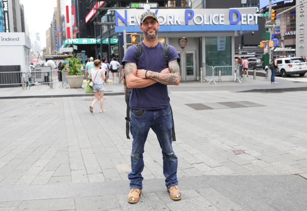 David Ramos poses in Times Square with a New York Police Department building behind him.
