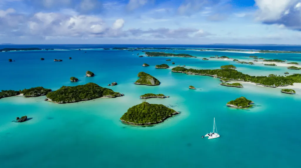 A drone image over Falaga Island in Fiji shows a catamaran anchored in turquoise water among many small, lush islands.