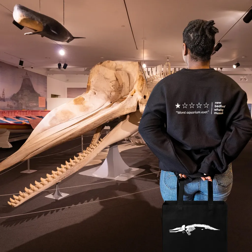 A person wearing a "Worst Aquarium Ever" sweatshirt and carrying a tote bag with a whale skeleton, stands in front of whale exhibits.