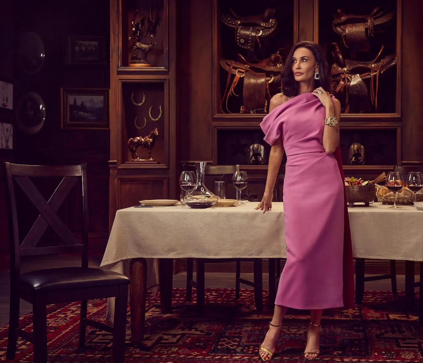Demi Moore in a pink dress, standing beside a dining table in a room decorated with Western-themed items.