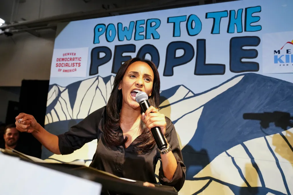 Melat Kiros speaking into a microphone with a backdrop that reads "POWER TO THE PEOPLE" and "DENVER DEMOCRATIC SOCIALISTS OF AMERICA".