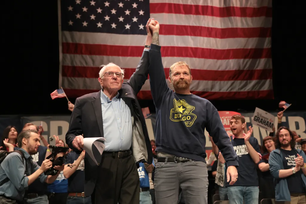 Bernie Sanders and Graham Platner raising their joined hands in front of an American flag.