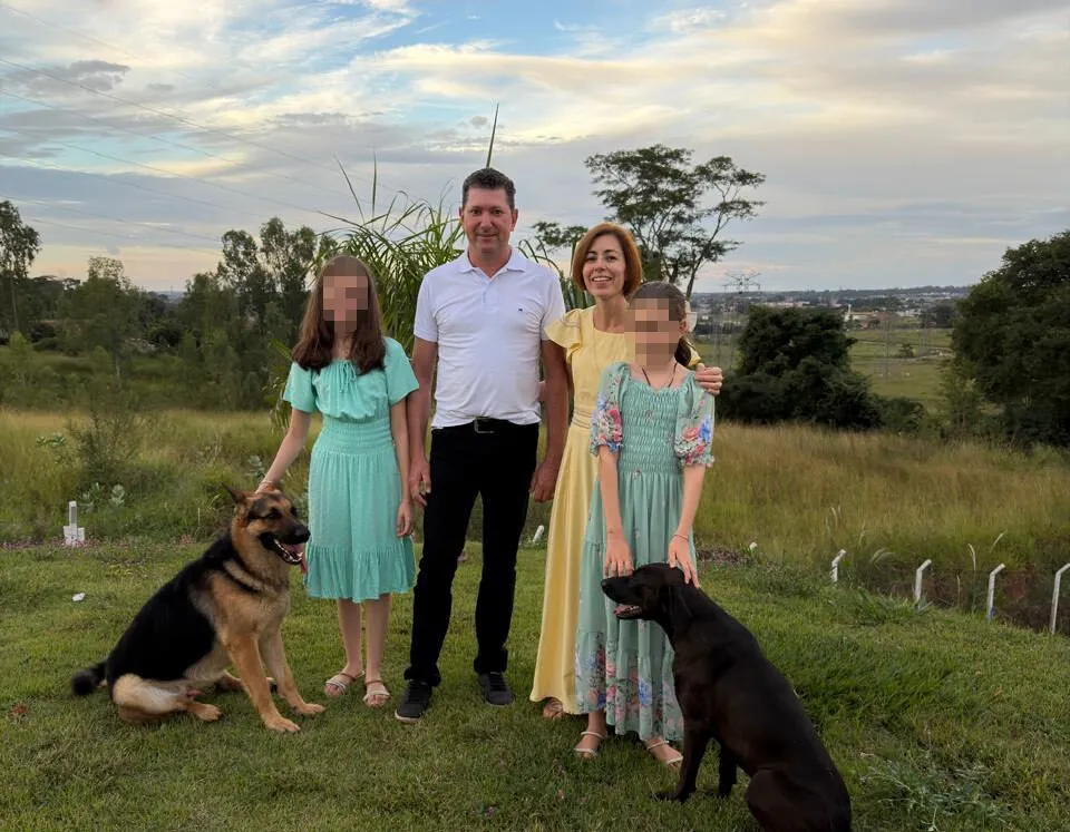The Denardi family, a couple with two daughters and two dogs, stands outdoors against a cloudy sky and green landscape.