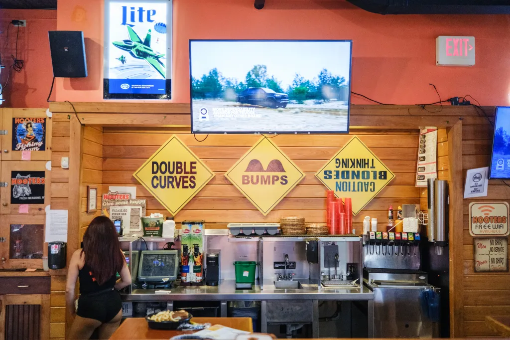 Interior of Hooters restaurant with a server at the counter, a TV playing an advertisement, and signs.