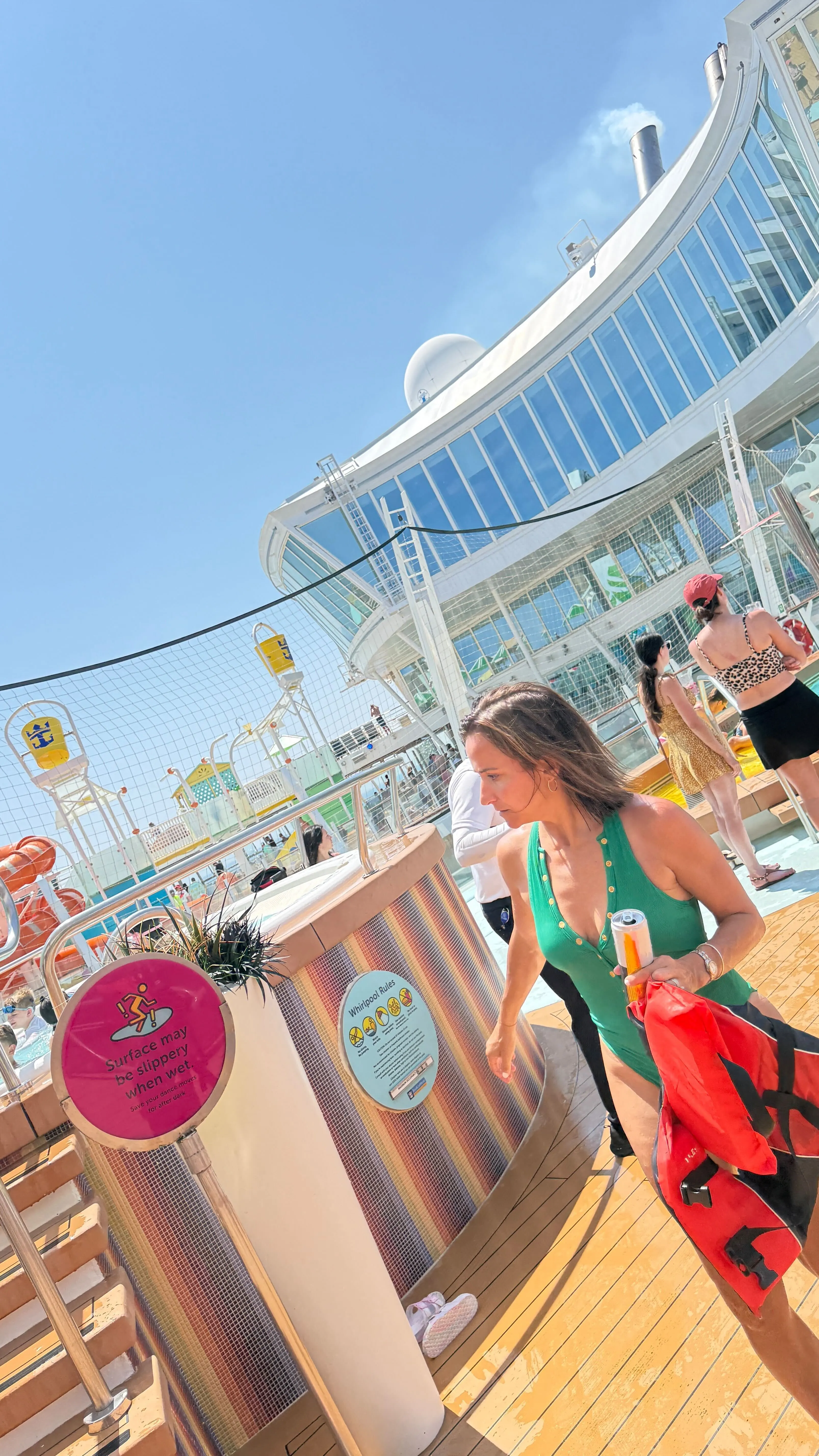 Dianna Russini walking on a cruise ship deck in a green swimsuit, holding a life vest and a drink.