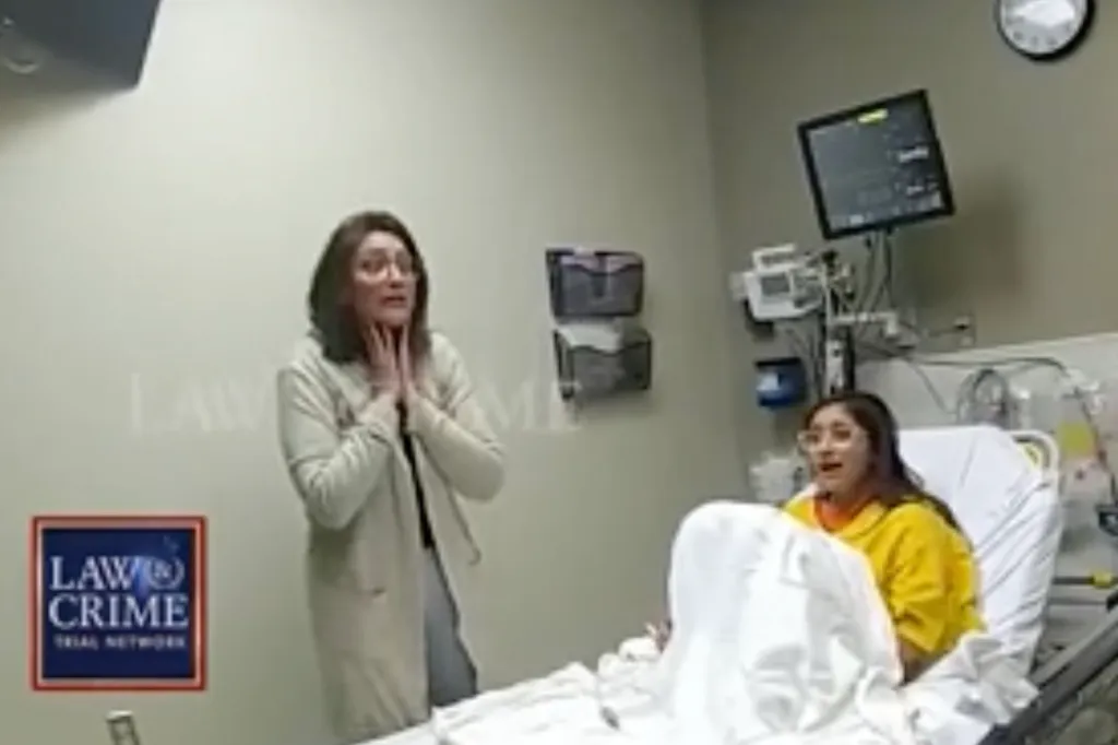 A woman in a hospital gown sits in a bed, looking shocked, while another woman stands beside her, holding her hands to her throat in distress.