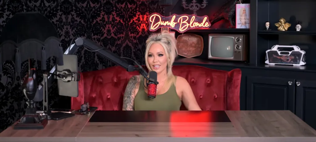 Bunnie XO seated in a red armchair at a desk with a microphone, in front of a wall with black damask wallpaper.