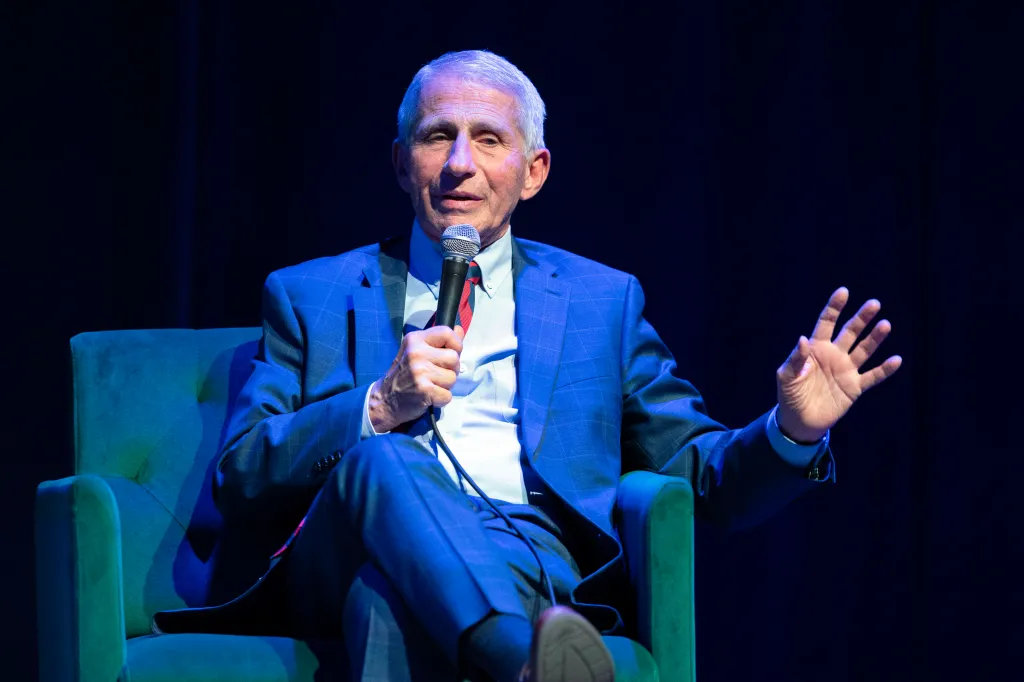 Dr. Anthony Fauci seated, speaking into a microphone, and gesturing with his left hand.