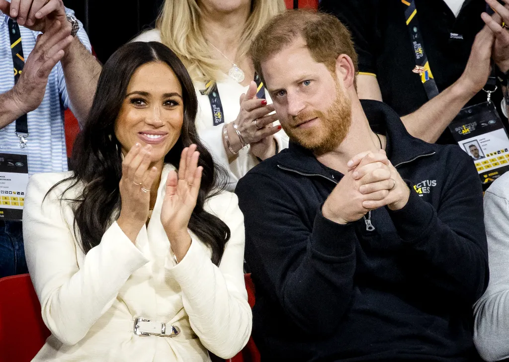 Meghan Markle clapping with a smiling face, while Prince Harry sits beside her with a neutral expression, both wearing Invictus Games passes.