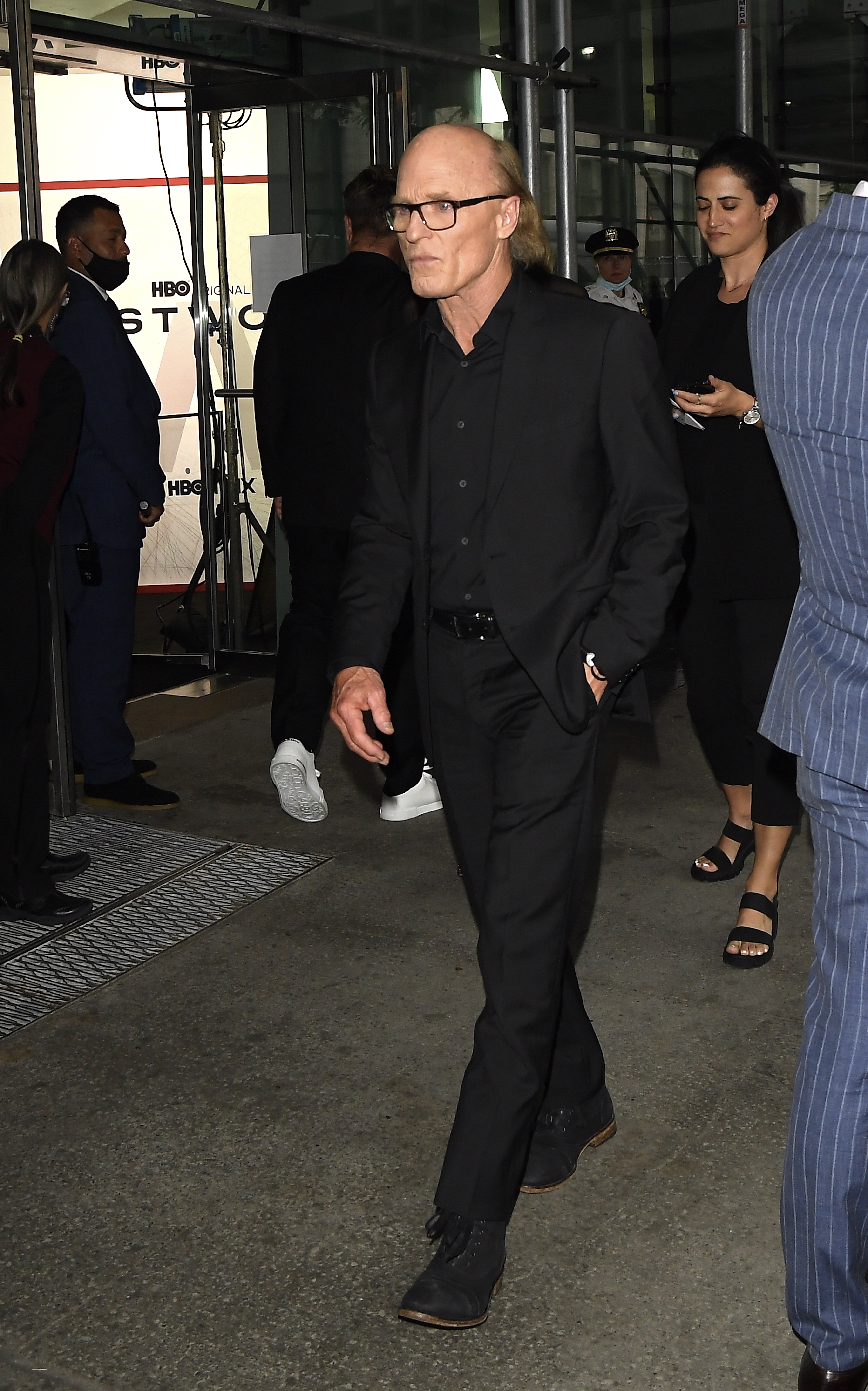 Ed Harris walking in an all-black suit and glasses, surrounded by people.