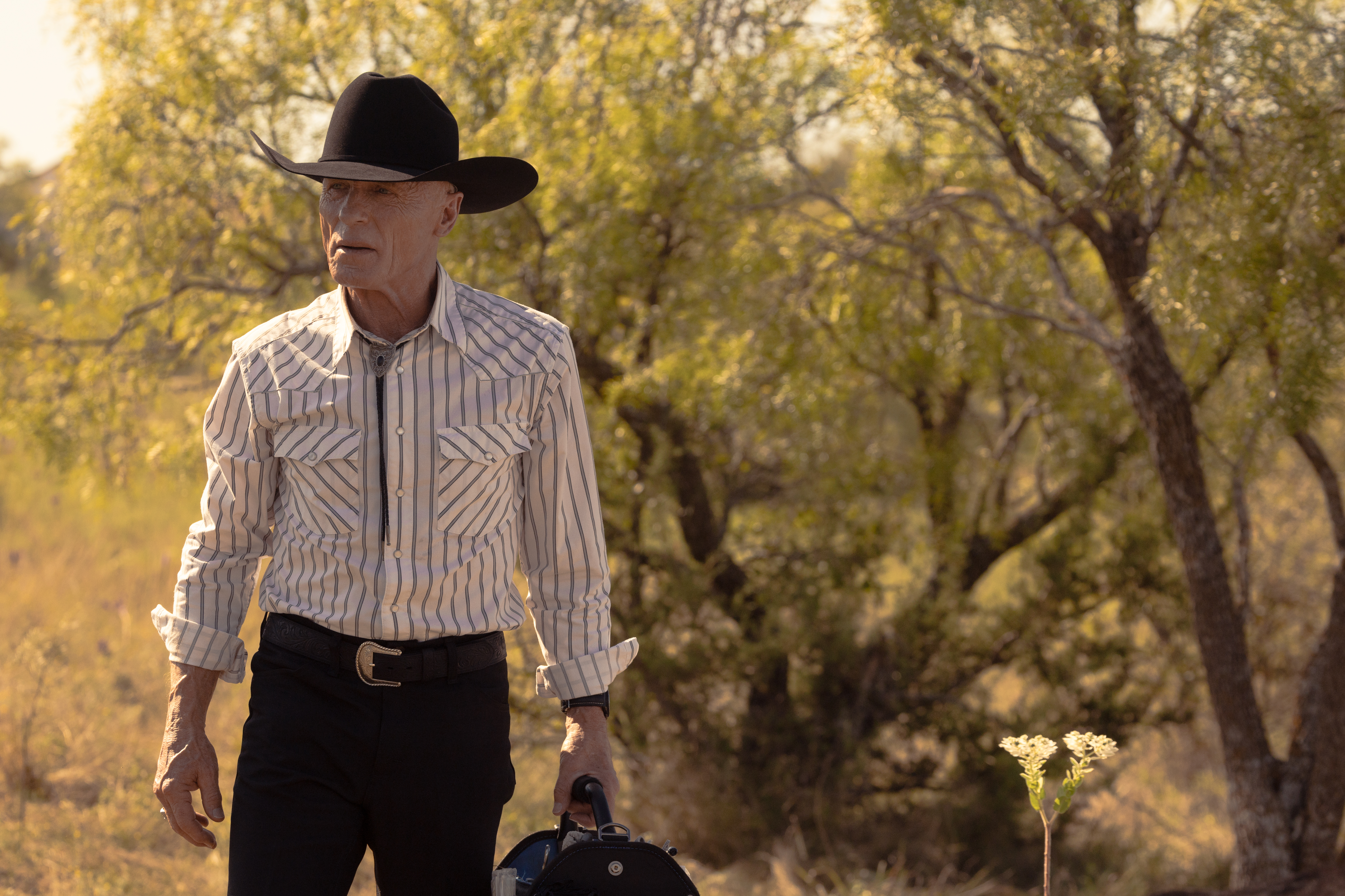 Ed Harris as Everett McKinney in a striped Western shirt and cowboy hat, holding a bag, with trees in the background.