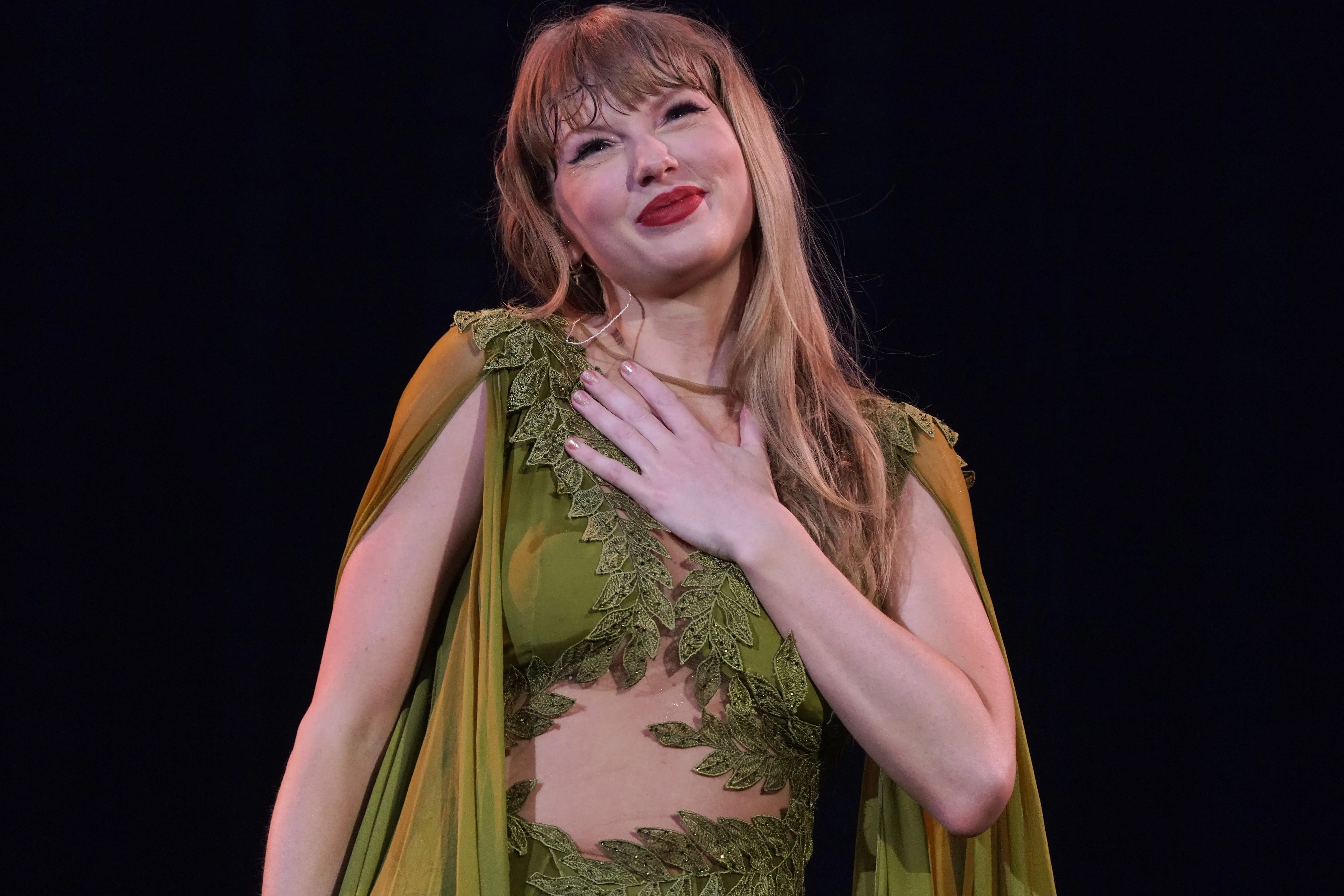 Taylor Swift performing onstage with her hand over her chest, wearing a sheer olive green gown with leaf-like embroidery.