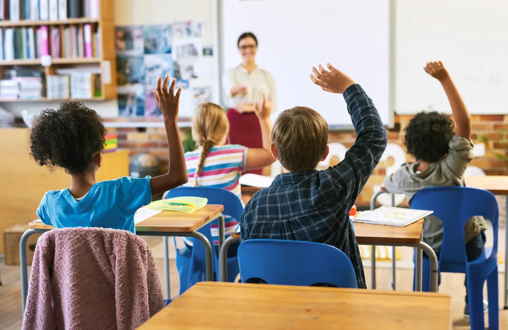 Four students in a classroom raise their hands to answer a question, while their teacher stands at the front.