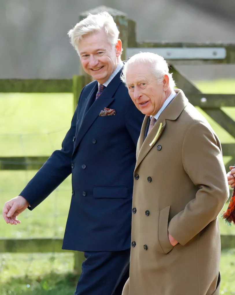 King Charles III, wearing a leek on his lapel, and Sir Clive Alderton walking outdoors.