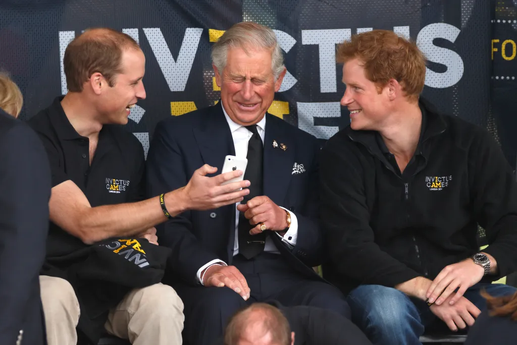 Prince William, in a black T-shirt and khaki pants, King Charles, in a navy blue suit, and Prince Harry, in a black jacket and blue jeans, laughing while looking at a phone during the Invictus Games.