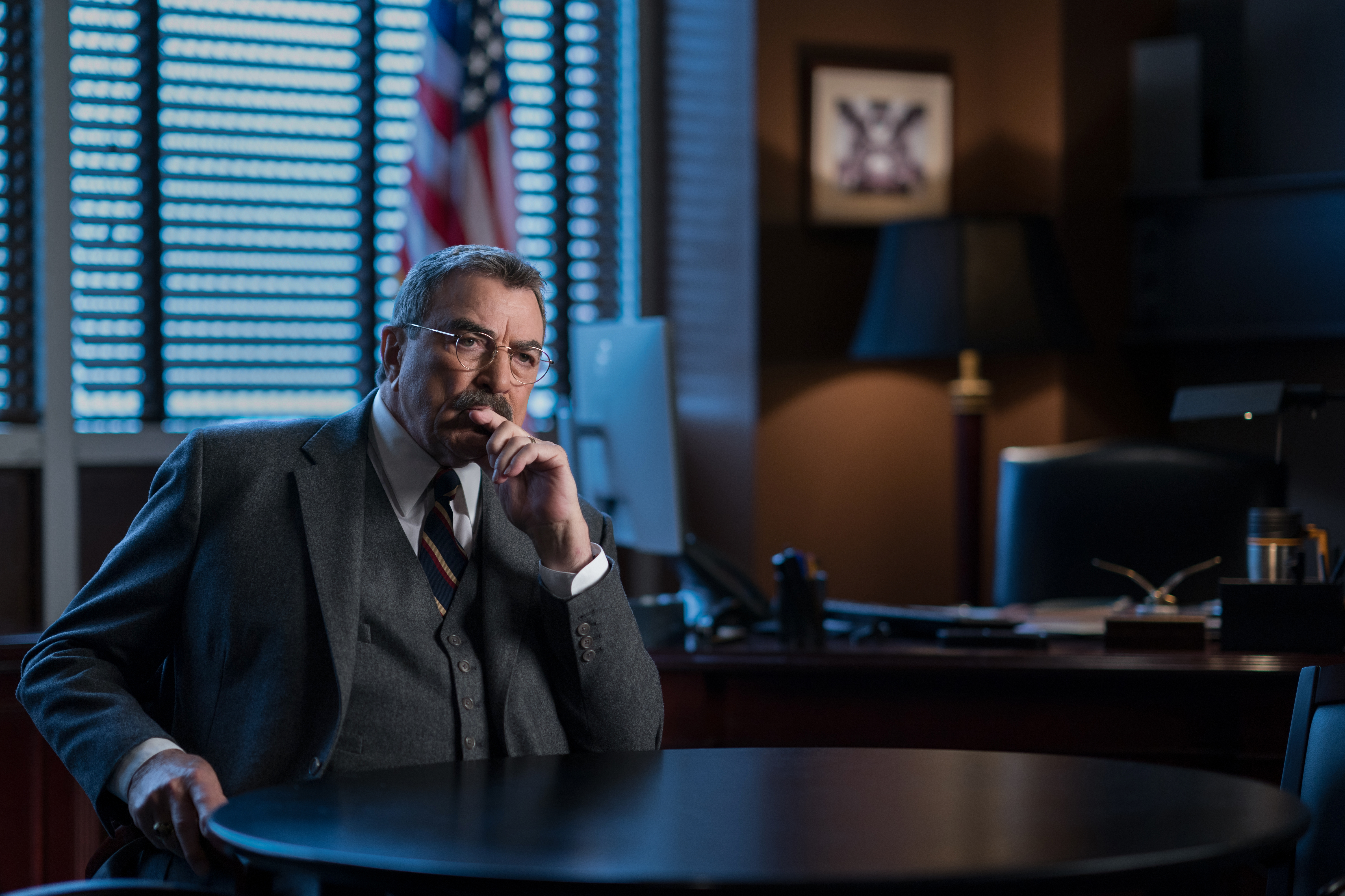 Tom Selleck as Frank Reagan from "Blue Bloods" sitting at a table with his hand on his chin.