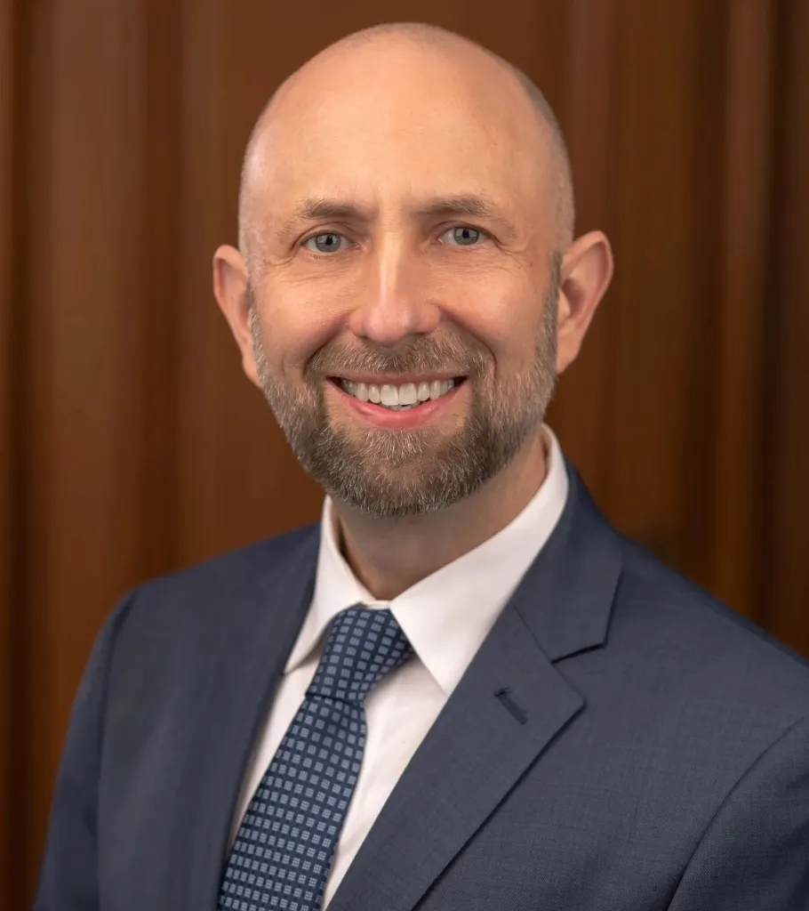 Headshot of Joel Engardio, a bald man with a beard, smiling and wearing a blue suit and patterned tie.