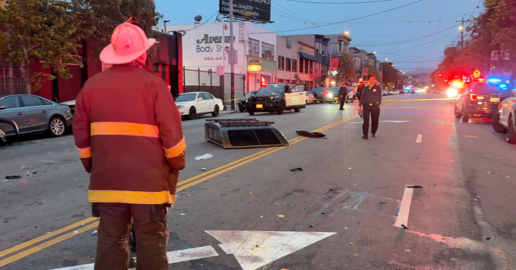 A firefighter stands in the foreground, facing away from the camera, as first responders assess the scene of a multi-vehicle collision with flashing lights in the background.