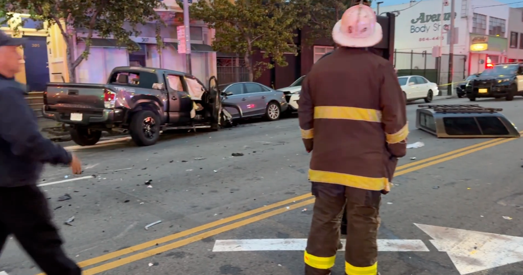 Multi-vehicle collision with a dark grey pickup truck and silver sedan, debris on the road, and emergency personnel present.