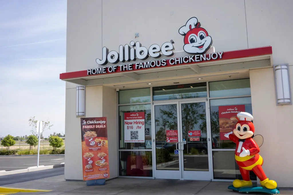 The entrance to a Jollibee restaurant in Delano, California, featuring its mascot "Jollibee" and signs for hiring and menu deals.