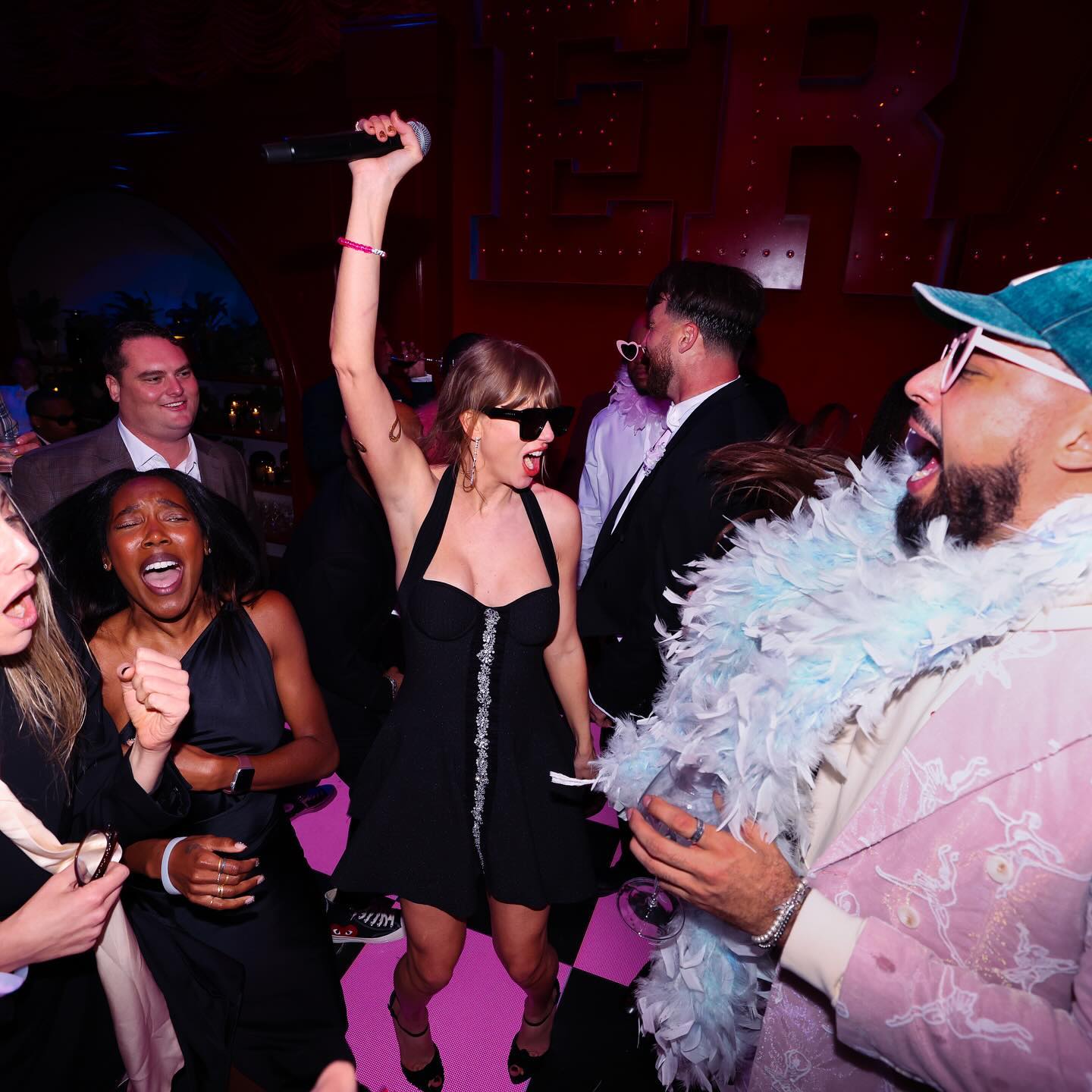 Taylor Swift singing into a microphone with her arm raised at a party, surrounded by other enthusiastic guests.
