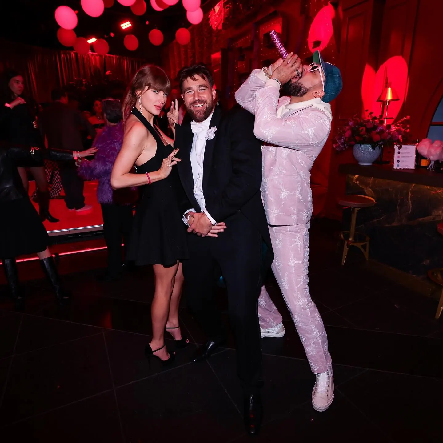 Taylor Swift, Travis Kelce, and Ross Travis posing at a party with red lighting.