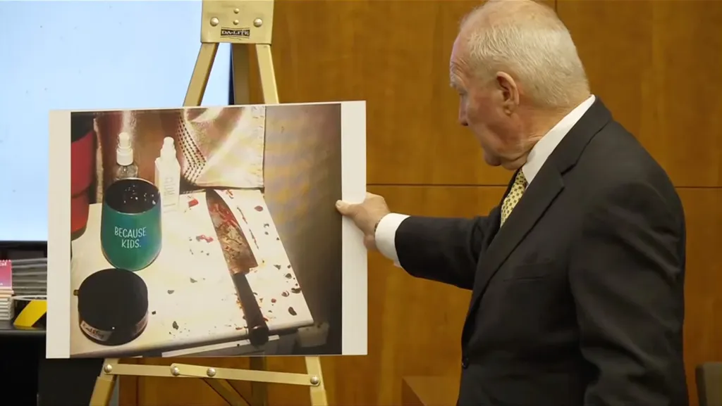 A man in a suit displays a crime scene photo showing a bloody knife, a cup that reads "BECAUSE KIDS.", and other items.