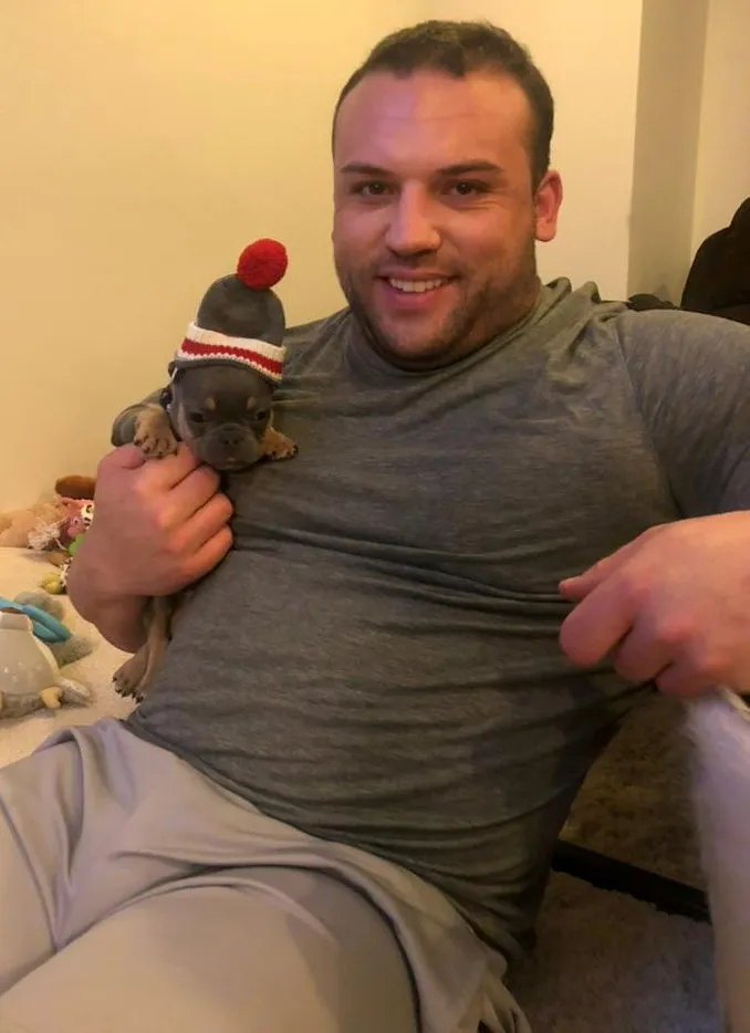 Nick Trigili smiles while holding a puppy wearing a knitted hat.