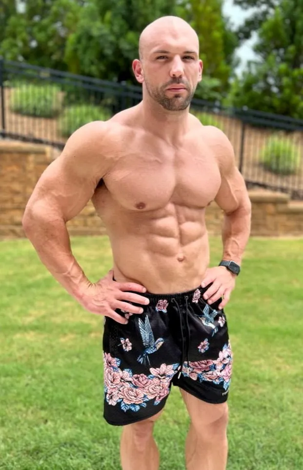 Nick Trigili, a muscular man with a shaved head and beard, wearing floral swim trunks.