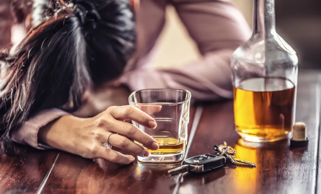 A woman in a pink shirt passed out on a table with a glass of whiskey, a bottle of whiskey, and car keys.