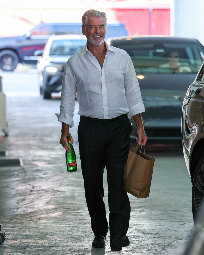 Pierce Brosnan walking with a bottle of water and a brown paper bag.