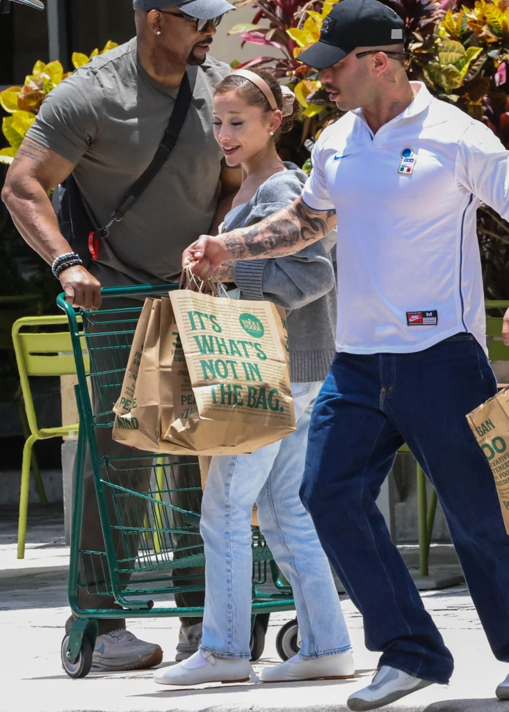 Ariana Grande in a gray hoodie, light blue jeans, and white shoes, holding Whole Foods grocery bags while walking with two men.