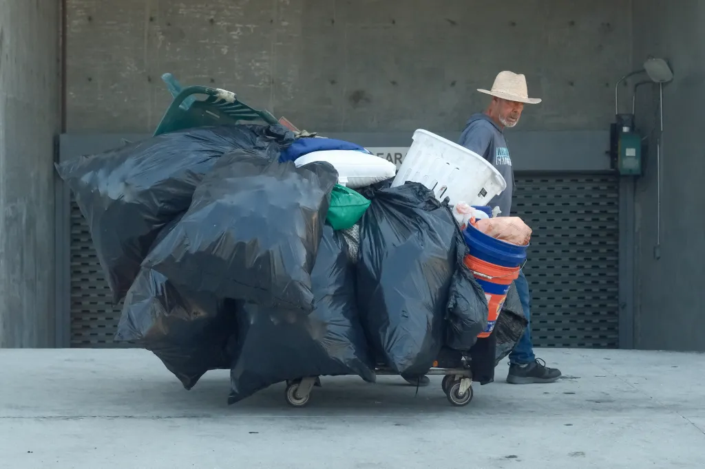 Man with a straw hat pushes a cart overflowing with black trash bags, pillows, and buckets.