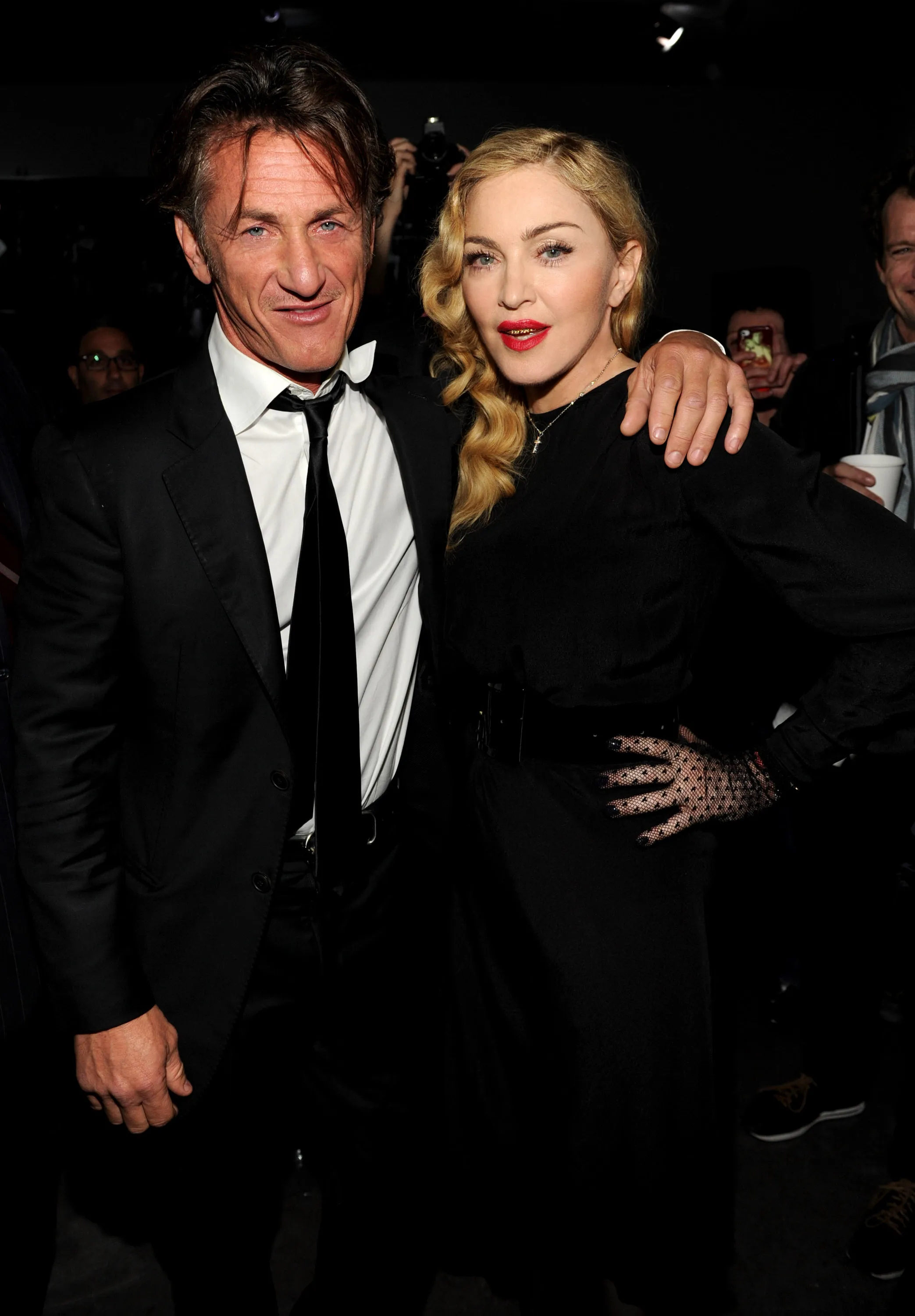Sean Penn, wearing a black suit and tie, and Madonna, in a black dress with red lipstick and a gold grill, pose together.