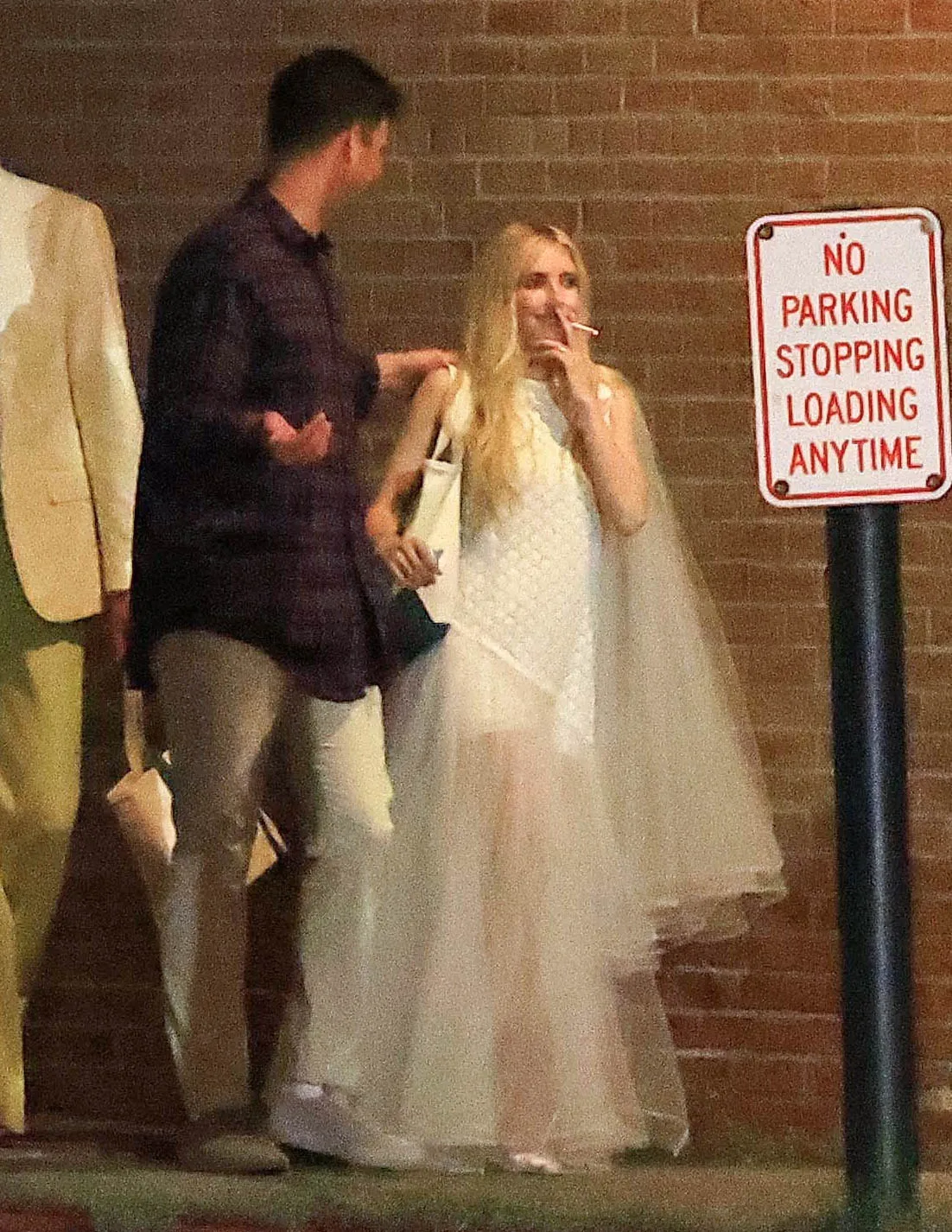 Emma Roberts in a white dress, smoking a cigarette, next to a man and a "NO PARKING" sign.