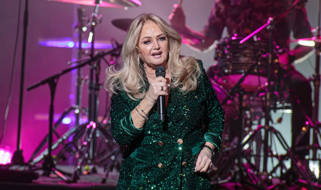Bonnie Tyler singing into a microphone on stage, wearing a sparkly green jacket.
