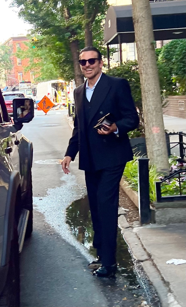 Bradley Cooper smiling in a suit and sunglasses, walking on a street with construction in the background.