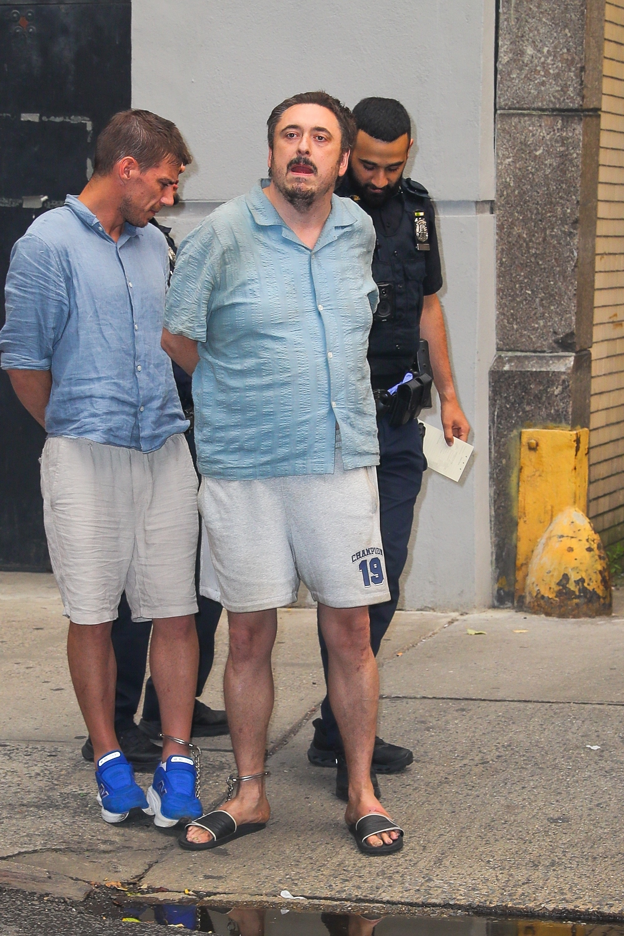 Andrew Truelove, the suspected Today Show intruder, is seen in shackles outside an NYPD precinct, accompanied by another individual and a police officer.