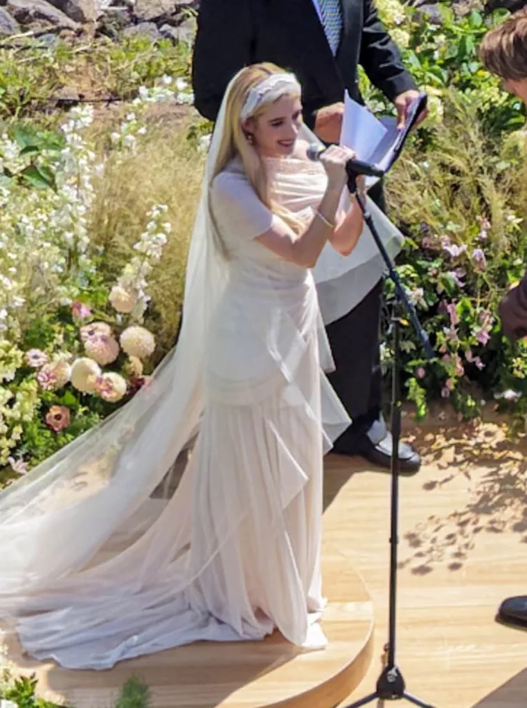 Emma Roberts at her wedding to Cody John, dressed in a white wedding gown with a veil and holding a microphone while smiling.